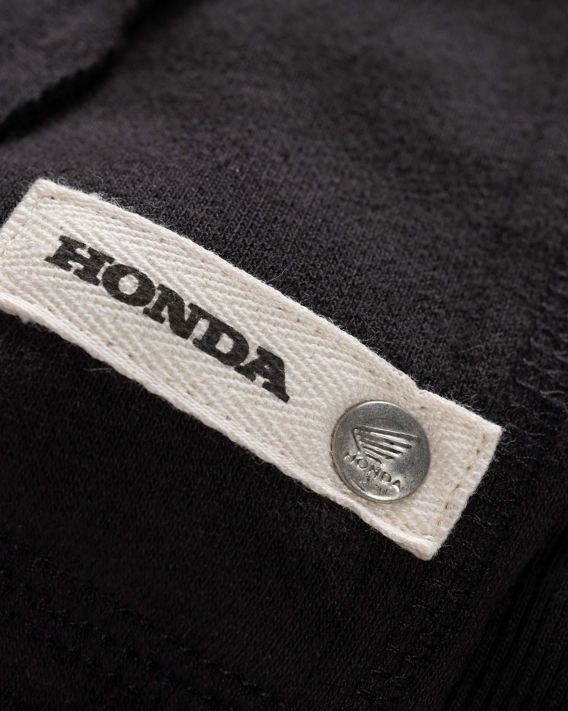 Beiges Webetikett mit Honda Schriftzug und silbernem Druckknopf am schwarzen Sunset Hoodie.